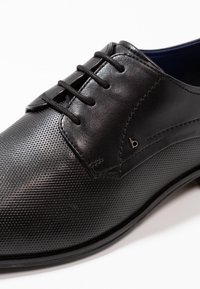Zapatos de cuero negro con un acabado texturizado, cierre con cordones y un pequeño emblema en el lateral. Acentos suaves en la parte superior.