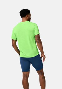 Camisa de manga corta de un tejido verde fluorescente, con una textura suave. Combinada con pantalones cortos ajustados de color azul oscuro. Tatuajes visibles en el brazo.