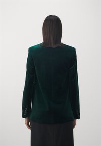 Grön sammetblazer med skräddarsydd passform, strukturerade axlar och knappade ärmar, sedd bakifrån mot en neutral bakgrund.