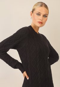 Czarny sweter robiony na drutach z teksturowanym wzorem rombów, z długimi rękawami i okrągłym dekoltem. Materiał wydaje się miękki i gruby.