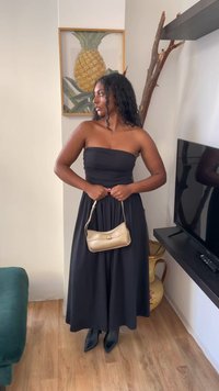 Svart strapless klänning med en slät textur, parat med en liten guldhandväska och knähöga svarta stövlar. Neutral bakgrund med dekoration.