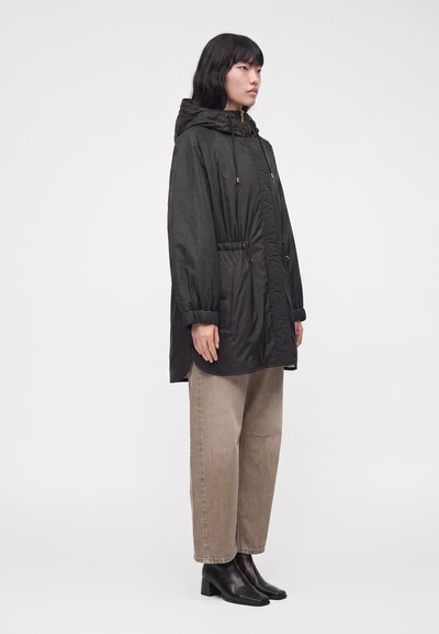 Personne portant un manteau à capuche noir avec cordons, un pantalon large beige et des bottines noires, debout de côté sur un fond uni.