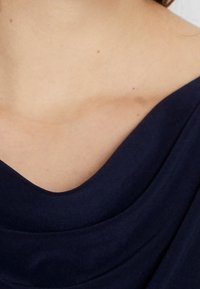 Tela drapeada de color azul marino con una textura suave. Muestra un escote ancho con contornos suaves. Primer plano de la zona del hombro con un tono de piel natural.