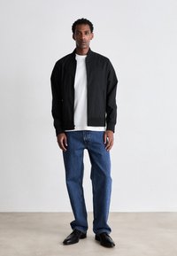 Giacca bomber nera con zip frontale, t-shirt bianca sotto, abbinata a jeans blu a gamba dritta e scarpe nere. Tessuto morbido e vestibilità su misura.