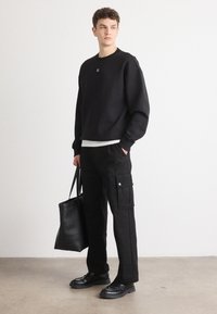 Sudadera negra con cuello redondo acanalado, combinada con pantalones cargo negros que tienen bolsillos laterales. El modelo sostiene una bolsa tote negra.