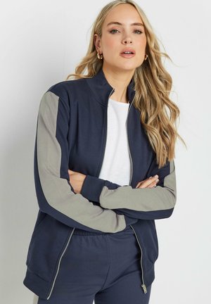 Marineblaues Zip-up-Jackett mit hellgrauen Ärmeln, gerippten Bündchen und Kragen. Der Stoff ist weich und hat eine glatte Oberfläche. Wird mit einem weißen Shirt getragen.