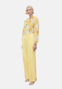 Bluză florală cu trandafiri galbeni pe un fundal alb, asortată cu pantaloni din in galbeni cu talie înaltă. Modelul stă pe un fundal simplu.
