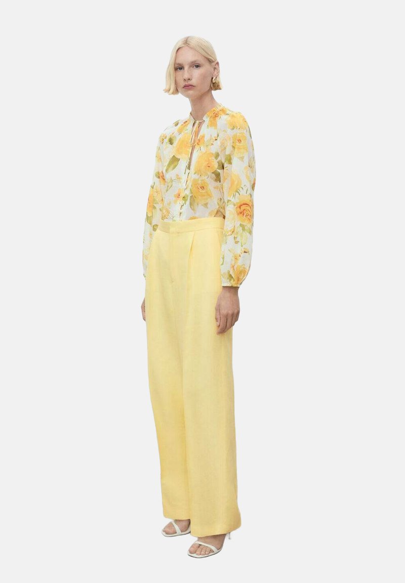 Bluză florală cu trandafiri galbeni pe un fundal alb, asortată cu pantaloni din in galbeni cu talie înaltă. Modelul stă pe un fundal simplu.