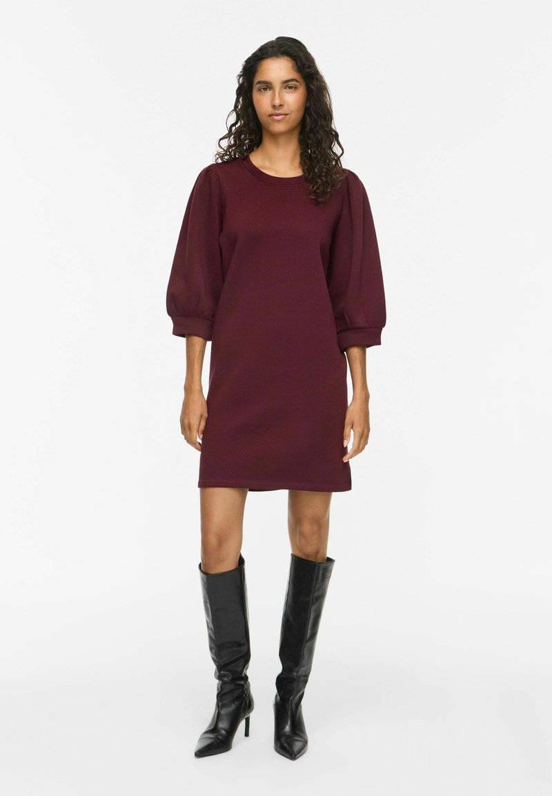 Femme aux longs cheveux bouclés portant une robe longue violette foncée aux genoux avec des manches bouffantes et des bottes noires à talons hauts montant jusqu'aux genoux, debout face à l'avant.