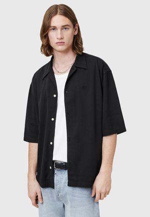Camisa negra de manga corta y botones con un corte relajado, que presenta un sutil logo bordado, combinada con una camiseta blanca de debajo y jeans claros.