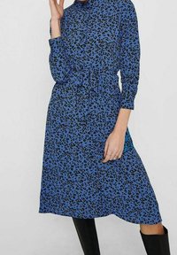 Abito camicia blu con stampa leopardata, chiusura con bottoni, maniche lunghe, vita cinchata e lunghezza fino al ginocchio. Abbinato a stivali neri al ginocchio.
