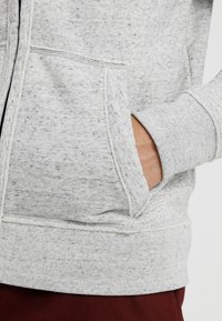 Sudadera gris salpicada con cremallera frontal, bolsillo tipo canguro, puños y dobladillo de canalé. El material parece suave y texturizado.