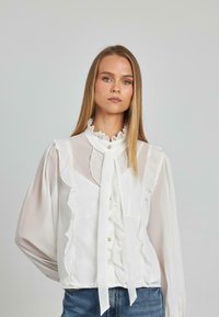 Weiße Seidenbluse mit Rüschendetails, hohem Kragen und Knopfverschluss. Lange Ärmel mit transparenter Textur. Kombiniert mit Jeans.