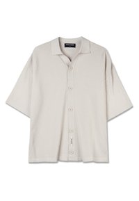 Camisa de manga corta color beige pálido con textura acanalada, cierre de botones en la parte frontal y un diseño de ajuste relajado. Presenta un cuello clásico y detalle de etiqueta en el lateral.