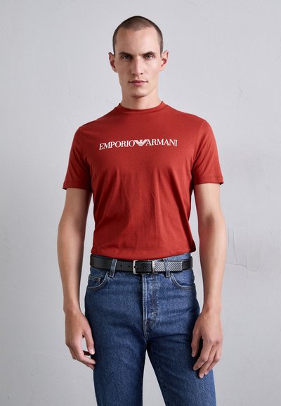 Tricou ro�șu din bumbac cu guler rotund și logo-ul "EMPORIO ARMANI" imprimat în alb. Asortat cu blugi albaștri și o curea neagră.