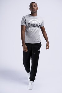 Gray Lonsdale t-shirt with black text, featuring a lion graphic. Paired with black jogger pants and white sneakers, standing on a light backdrop.