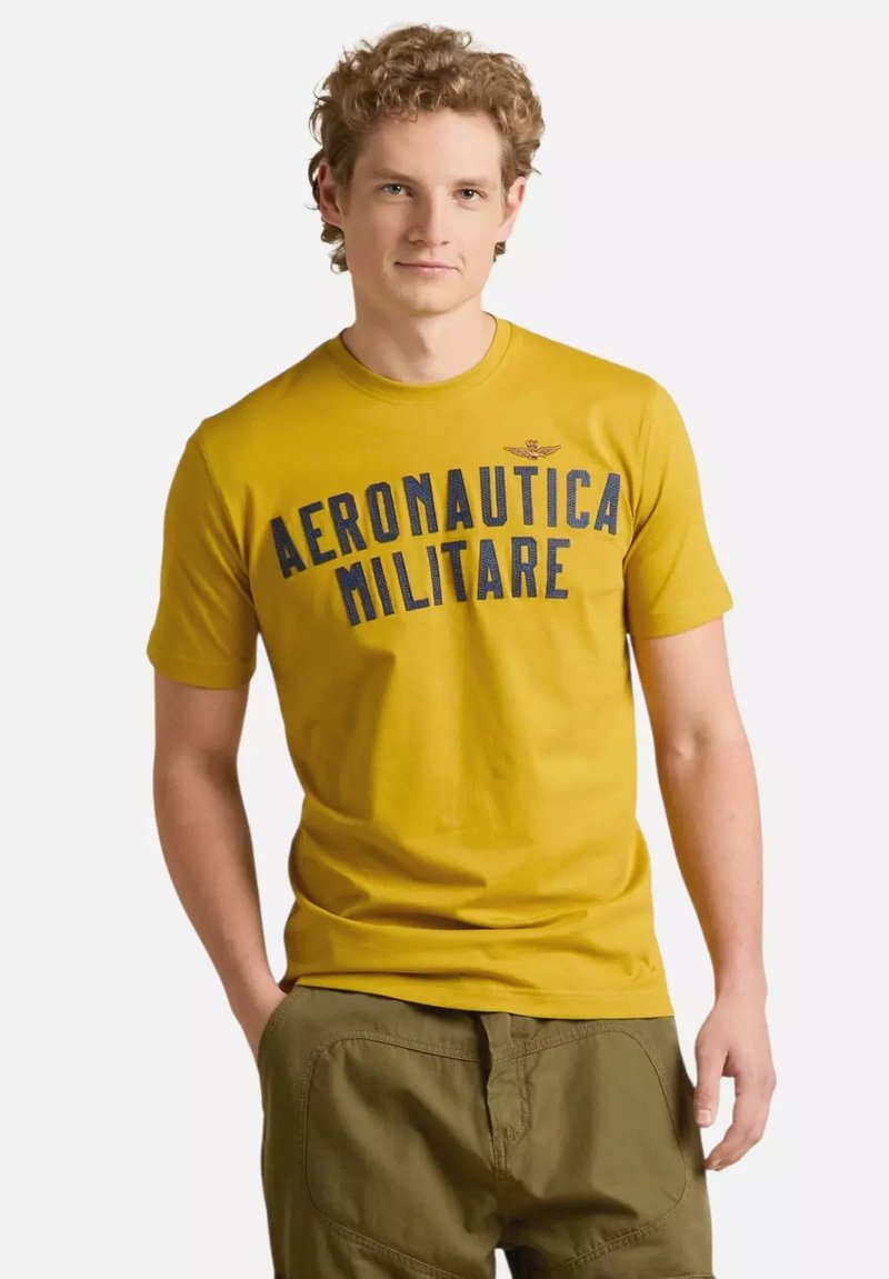T-shirt in cotone giallo con grande scritta blu "AERONAUTICA MILITARE", maniche corte e colletto girocollo, abbinato a pantaloni verde oliva.