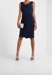 Vestido de gala sin mangas en color navy con un escote drapeado, silueta hasta la rodilla y detalle de envoltura. Combinado con un bolso de mano plateado y zapatos metálicos de tiras.