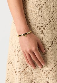 Goldene Armspange mit glatter Oberfläche und einem herzförmigen Anhänger, der einen kleinen Stein enthält. Tragend an einer Hand gegen ein gehäkeltes Kleid.
