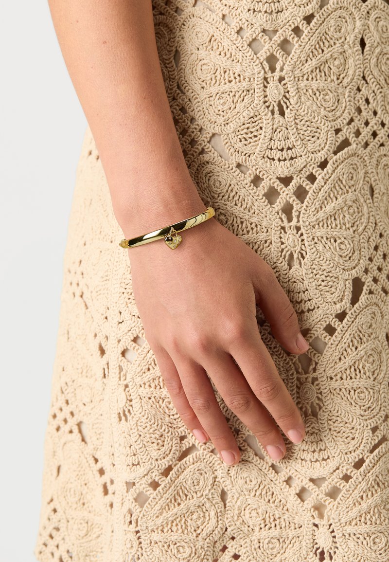 Goldene Armspange mit glatter Oberfläche und einem herzförmigen Anhänger, der einen kleinen Stein enthält. Tragend an einer Hand gegen ein gehäkeltes Kleid.