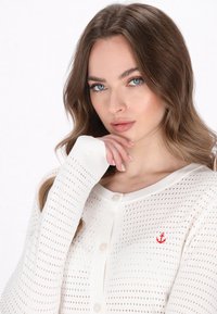 Cardigan bianco lavorato a maglia con un design perforato, caratterizzato da un collo rotondo, chiusura con bottoni e un piccolo emblema di un'ancora rossa sul lato sinistro del petto.