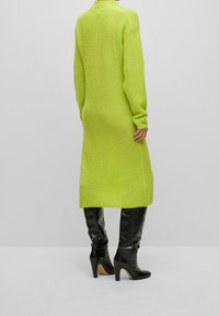 Longue robe pull tricotée de couleur vert citron vif, avec manches longues et col montant. Associée à des bottes noires à talons montant jusqu'au genou.