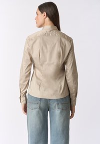 Camicia beige aderente con colletto, polsini con bottoni e pince posteriori, abbinata a jeans a gamba larga blu chiaro. Texture liscia, design casual.