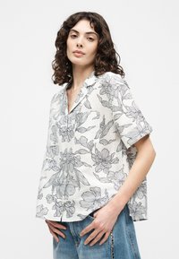 Γυναίκα με σγουρά μαλλιά που φοράει λευκό πουκάμισο με κοντά μανίκια και μαύρο φλοράλ σχέδιο, καθώς και μπλε τζιν, στέκεται μπροστά σε απλό φόντο.