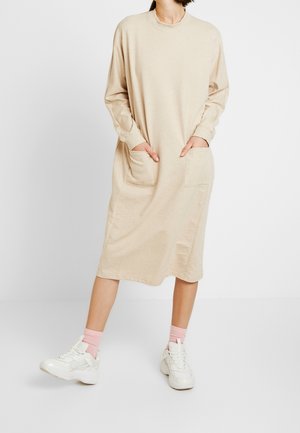 Beżowa bawełniana sukienka z długimi rękawami i dużymi przednimi kieszeniami, długością do kolan, zestawiona z białymi trampkami i różowymi skarpetkami za kostkę.