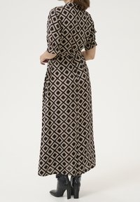 Robe maxi noire avec un motif de diamants en beige et blanc. Comprend des manches courtes roulées et une taille ceinturée. Portée avec des bottes à talons noires.