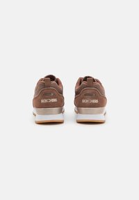 Brown suede sneakers with textured fabric, logo detailing, and a contrasting white midsole and outsole. Rear pull tabs for easy wear.