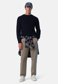 Suéter de punto acanalado marino, pantalones beige, camisa de cuadros atada a la cintura y gorra azul. Las zapatillas completan el look casual.