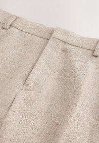 Tissu beige tissé, texturé avec un motif subtil. Présente une ceinture plate avec passants pour ceinture et des plis nets à l'avant.