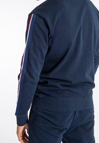 Mörkblå sweatshirt med rund halsringning, som har vertikala ränder i rött och vitt längs ärmarna. Mjuk textur, elastiska manschetter och nedre fåll.