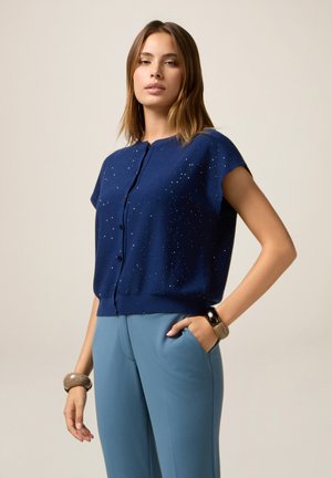 Junge Frau steht mit der Hand in der Tasche ihrer blauen Hose und trägt ein dunkelblaues Kurzarmhemd mit Knöpfen, das kleine weiße Punkte hat, sowie Armbänder.