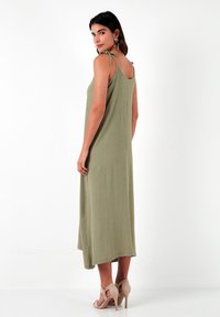 Olijfgroene maxi-jurk met dunne banden, gestructureerde stof, verstelbare strikdetails op de schouders en een asymmetrische zoom.