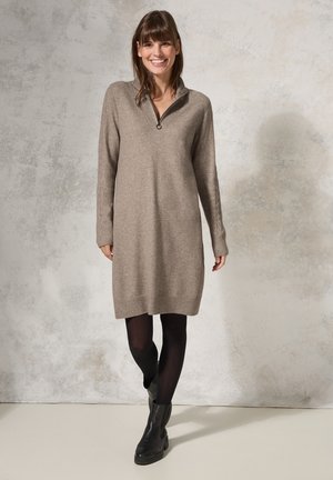 Beige stickad klänning med halvmatchande krage, ribbade ärmslut och kant, kombinerad med svarta strumpbyxor och ankelskoh. 