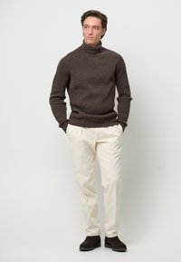 Brauner gerippter Rollkragenpullover mit taillierter Passform, kombiniert mit hellcremefarbenen Hosen und dunkelbraunen Wildlederschuhen.