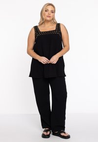 Zwart tweedelig outfit met een losse, gestructureerde tuniek met een vierkante halslijn en gouden stipaccenten, gecombineerd met bijpassende wijde broek.