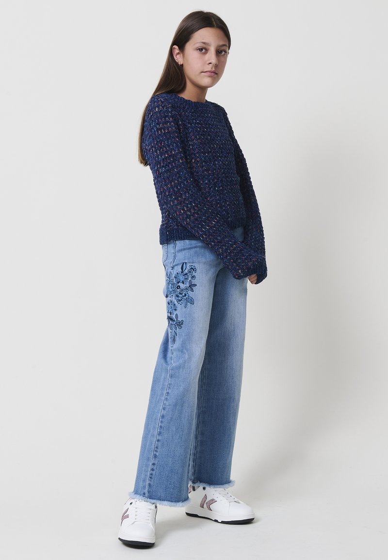 Ragazza in piedi con le braccia rilassate, indossa un maglione testurizzato blu scuro, jeans azzurri con ricami floreali e sneakers bianche.