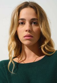Pull vert surdimensionné en tissu tricoté doux, avec un col large. Des cheveux longs et ondulés blonds et un collier en or complètent le look.