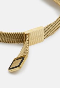 Pulseira de malha dourada com um fecho retangular apresentando riscas gravadas e o logótipo "GUESS". Acabamento texturizado com um detalhe em preto.
