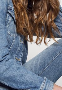 Chaqueta de mezclilla azul con un diseño clásico, que presenta un lavado claro y detalles deshilachados. Los jeans de mezclilla azul complementan la parte superior con un estilo ajustado.