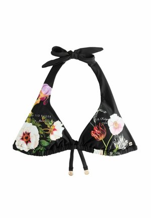 Frau, die ihre Haare richtet und einen schwarzen Blumen-Bikini trägt, steht am felsigen Ufer mit Ozeanwellen im Hintergrund.