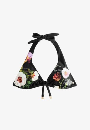 Femeie care își aranjează părul, purtând un bikini negru cu imprimeu floral, stând pe o țărm stâncos, cu valuri oceanice în fundal.