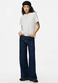 Camiseta blanca con lunaritos y mangas cortas, combinada con unos pantalones vaqueros de mezclilla oscura de pierna ancha. El modelo está de pie con las manos en los bolsillos, luciendo una boina negra.