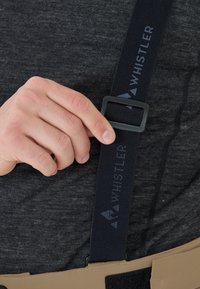 Schwarze verstellbare Hosenträger mit dem "WHISTLER"-Logo, einer mattschwarzen Klemme und einem strukturierten Riemen, der an einem sandfarbenen Kleidungsstück befestigt ist.