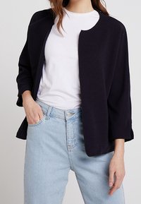 Svart ribbad cardigan med rund halsringning, trekvartslånga ärmar, öppen framsida och två sidofickor, bärs över en vit skjorta och ljusa jeans.