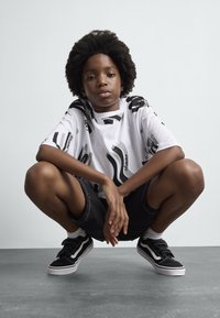 White t-shirt with black abstract patterns, black shorts, black and white Velcro sneakers, and a textured afro hairstyle.