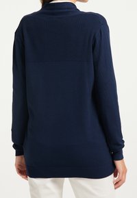 Marinblå tröja med ribbad struktur på övre delen av ryggen, långa ärmar och rak fåll. Mjukt stickat material, halvfigursydd design.